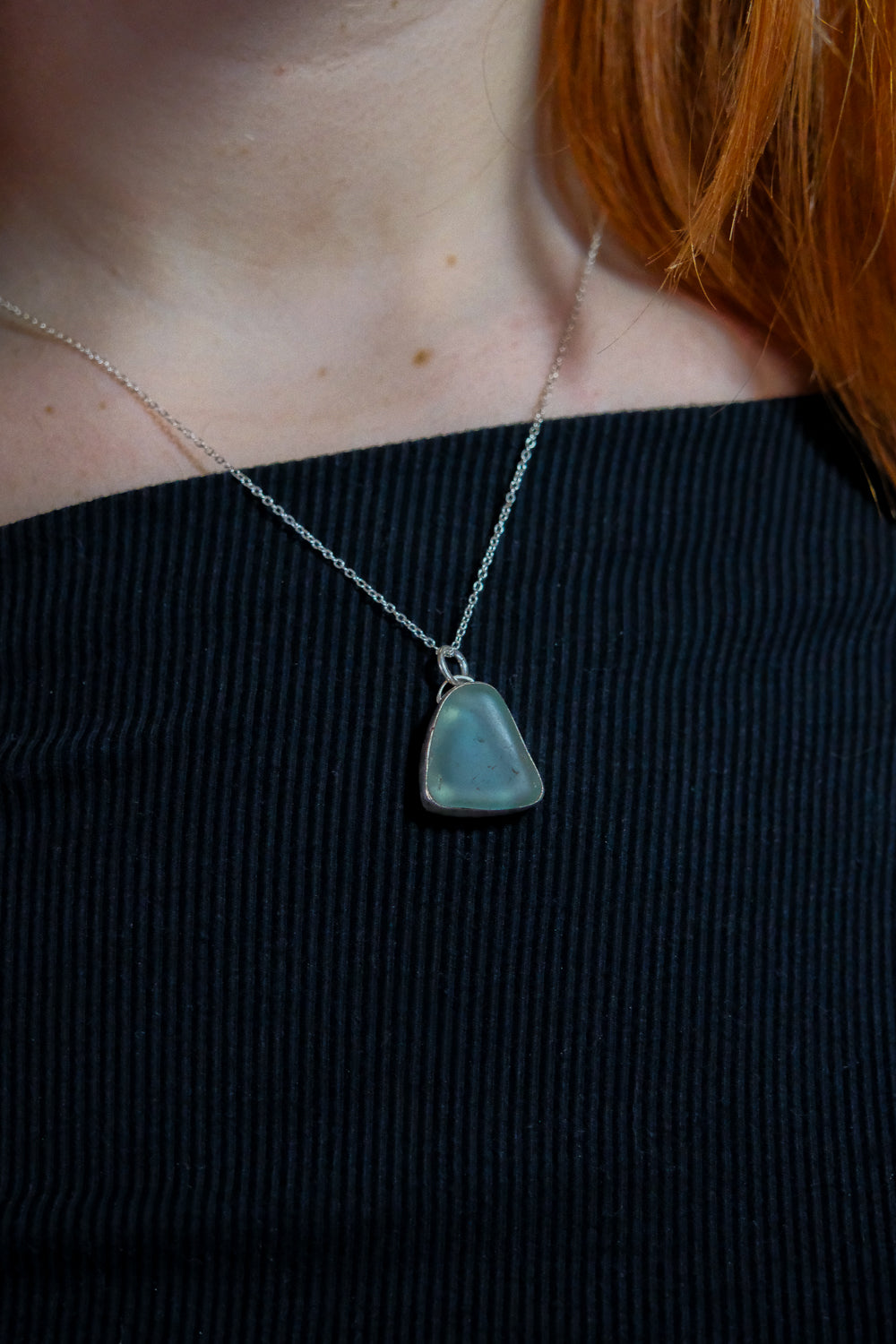 Necklace with a blue pendant worn by a person with red hair, against a black background
