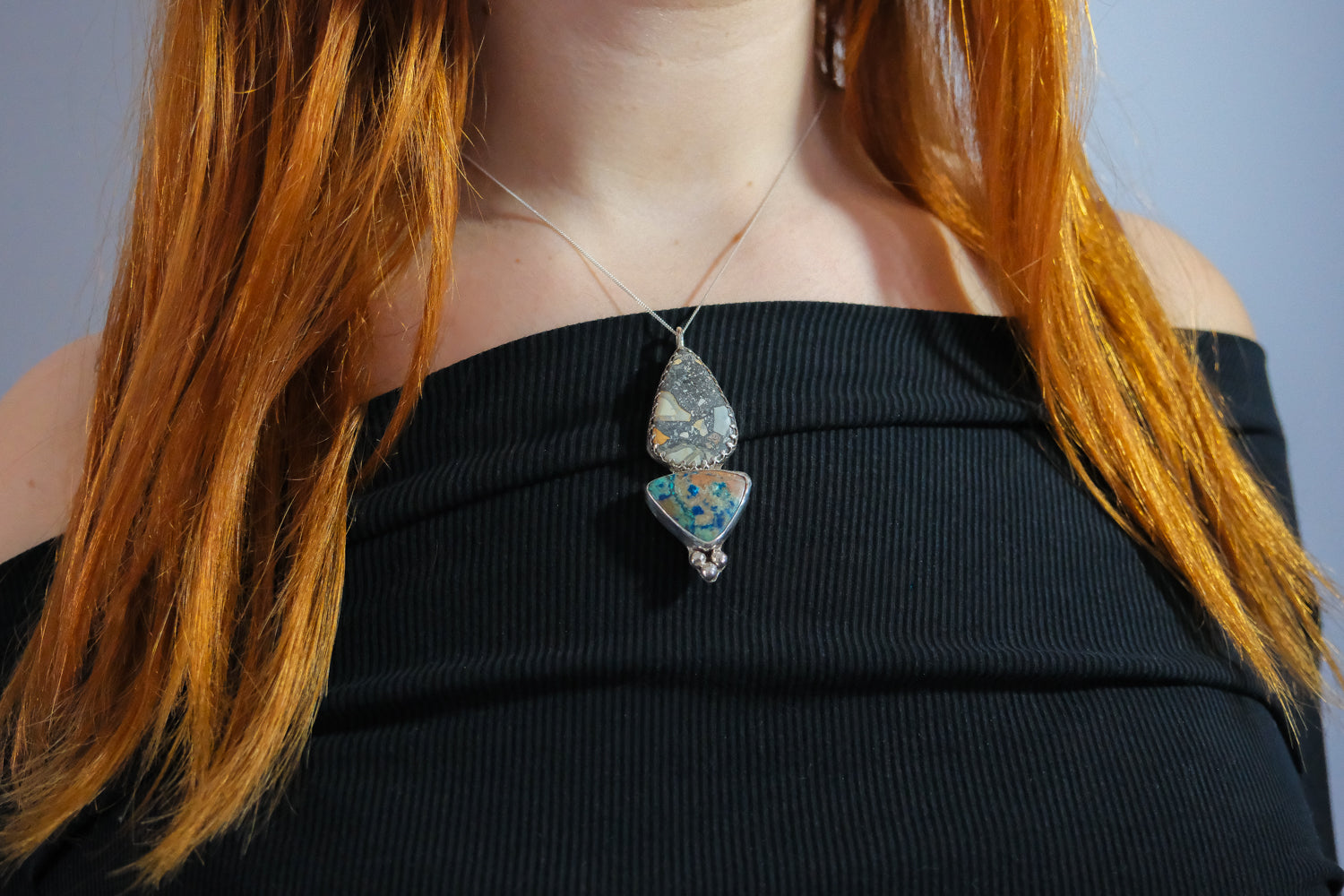 Person wearing a black top with a necklace featuring a blue pendant against a gray background