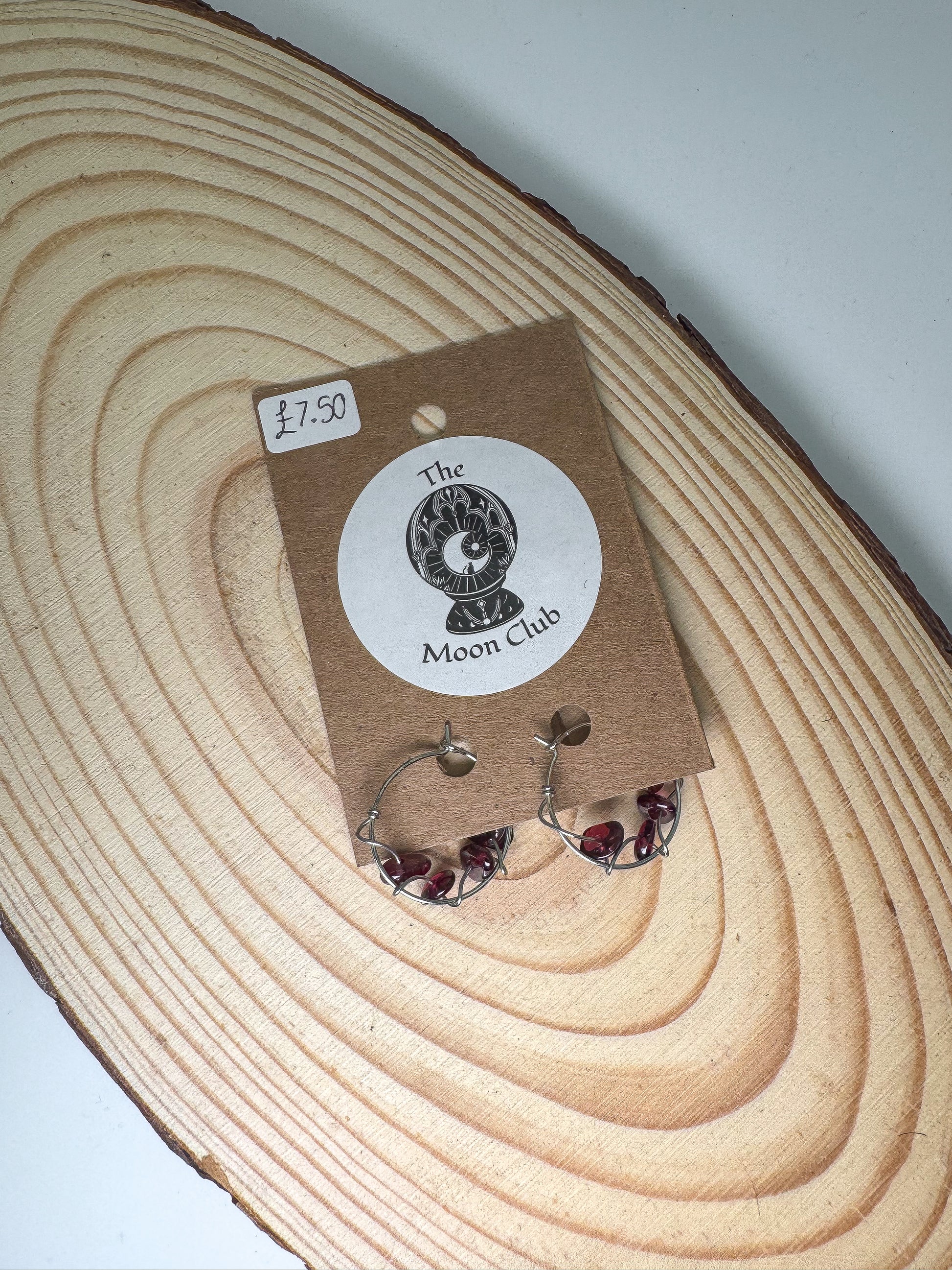 Pair of garnet earrings on a 'The Moon Club' branded card with a price tag, placed on a wooden surface.